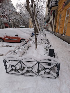 Snow-covered Budapest street with iron fences, parked cars, bare trees, and yellow building facade