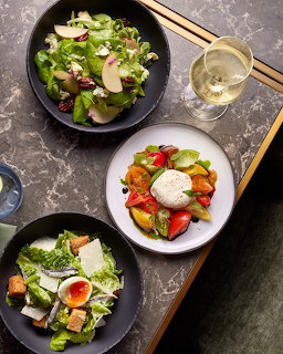 Three salad bowls with fresh greens, tomatoes, eggs, and vegetables on dark marble table with chopsticks