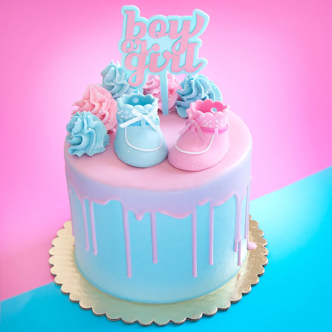 Pastel pink and blue drip cake with baby shoes, frosting swirls, and a topper