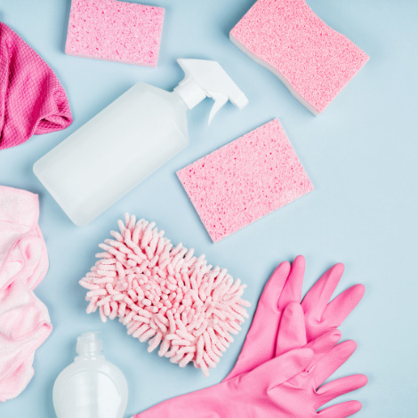 Pink cleaning supplies flatlay with sponges, rubber gloves, spray bottle, and cloths on a blue background