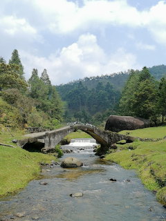 Old stone arch bridge over a shallow stream in a green mountain valley with large boulders