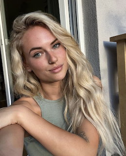 Portrait of a smiling blonde woman with wavy hair wearing a mint sleeveless top by a window