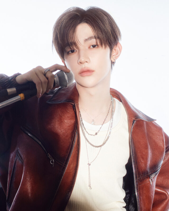 Young male K-pop singer holding a microphone in a brown leather jacket with layered necklaces