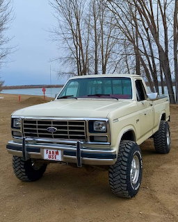 White lifted Ford pickup truck on beach with bare trees and lake background