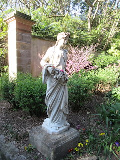 White classical female statue holding flowers on a stone pedestal in a lush garden setting