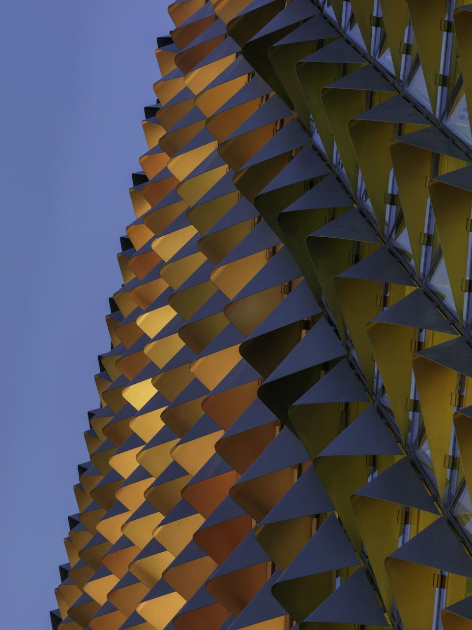 Modern building facade with repeating angular geometric panels in gold and gray against blue sky