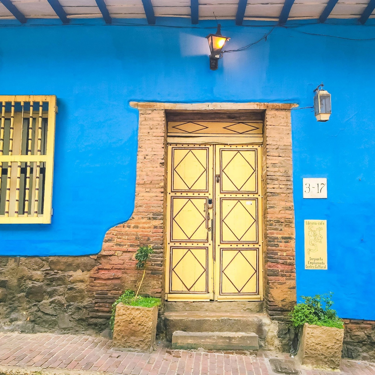 Bright blue wall with a cream double door, brick trim, window bars, lamps, and potted plants