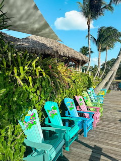Row of colorful Adirondack chairs on a wooden deck with palm trees and thatched roof in background