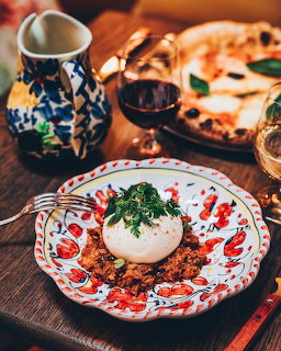 Burrata cheese with herbs on red-patterned plate, wine glass and pizza nearby on wooden table