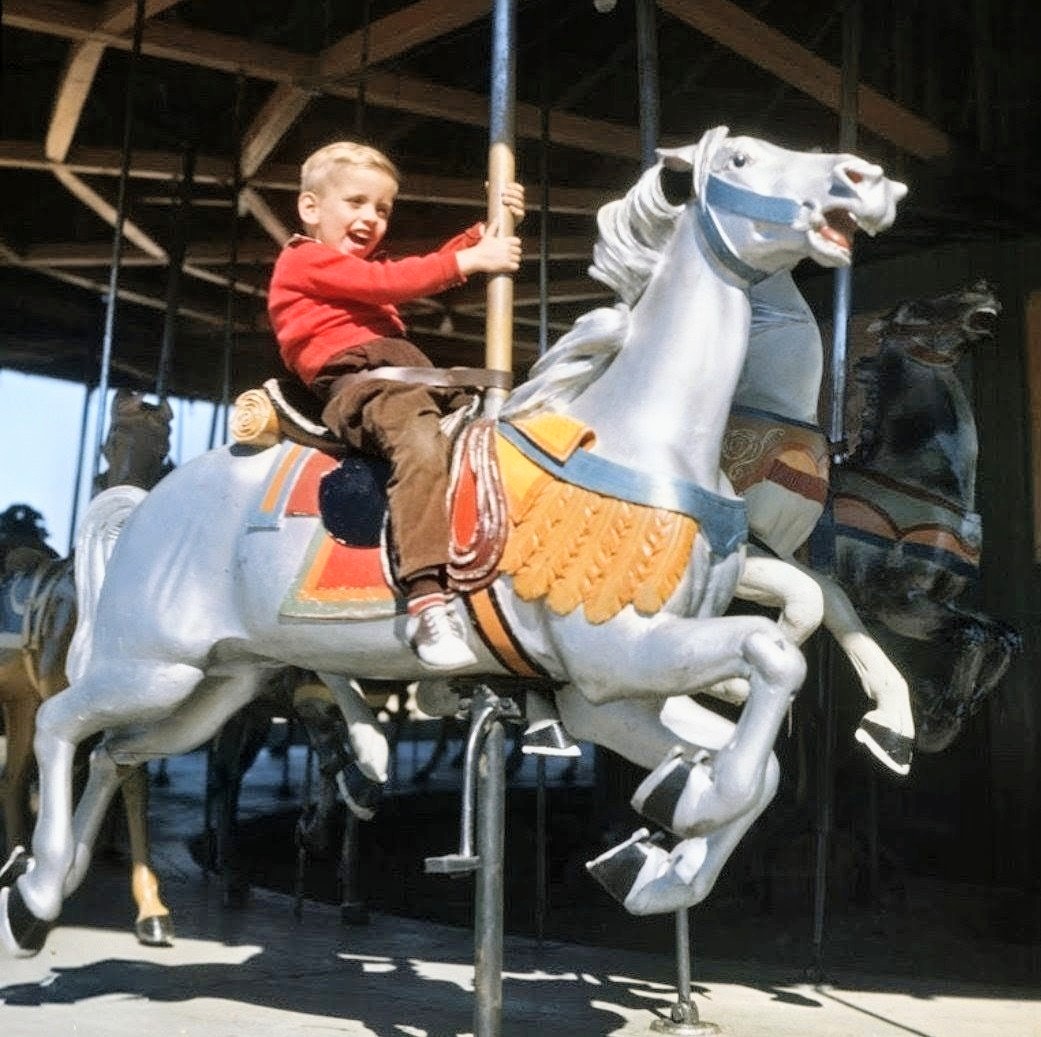 Young boy in red jacket smiling while riding a white carousel horse