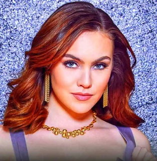 Close-up portrait of a woman with auburn hair, blue eyes, and gold jewelry against a blue background