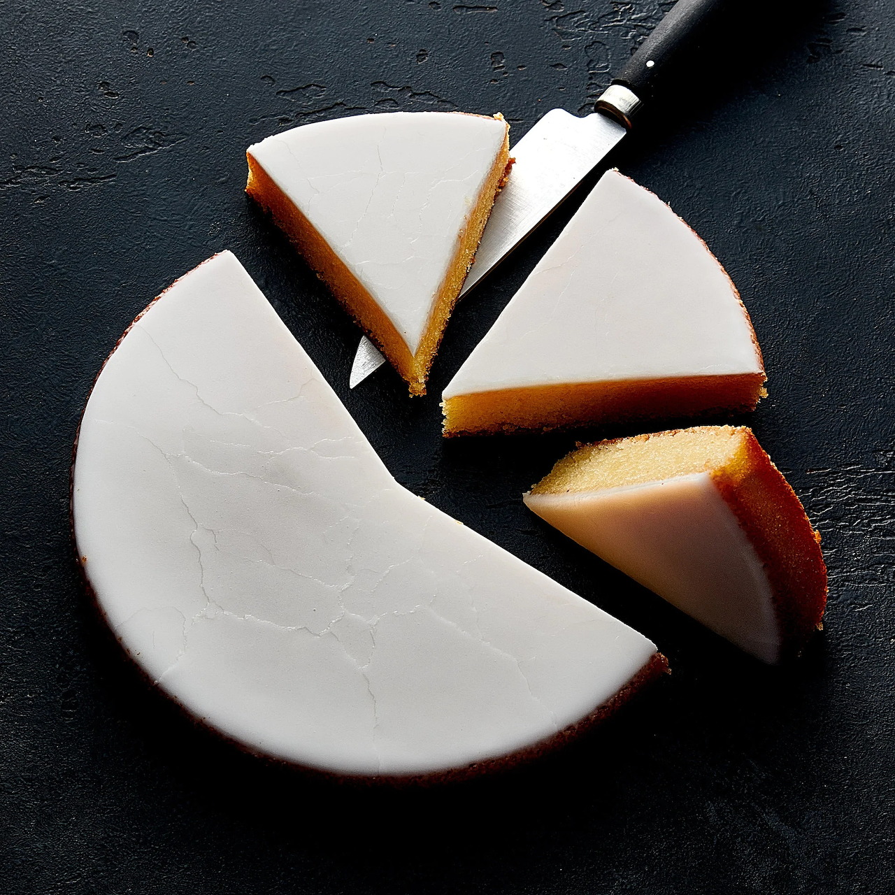 Round white glazed cake with slices and a cake server on a dark background