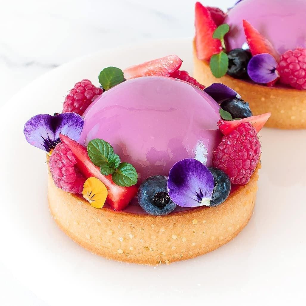 Two berry tarts with shiny pink glaze and colorful fresh fruit toppings on white marble surface
