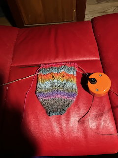 Colorful knitted fabric on knitting needles lying on a red leather couch with an orange tape measure