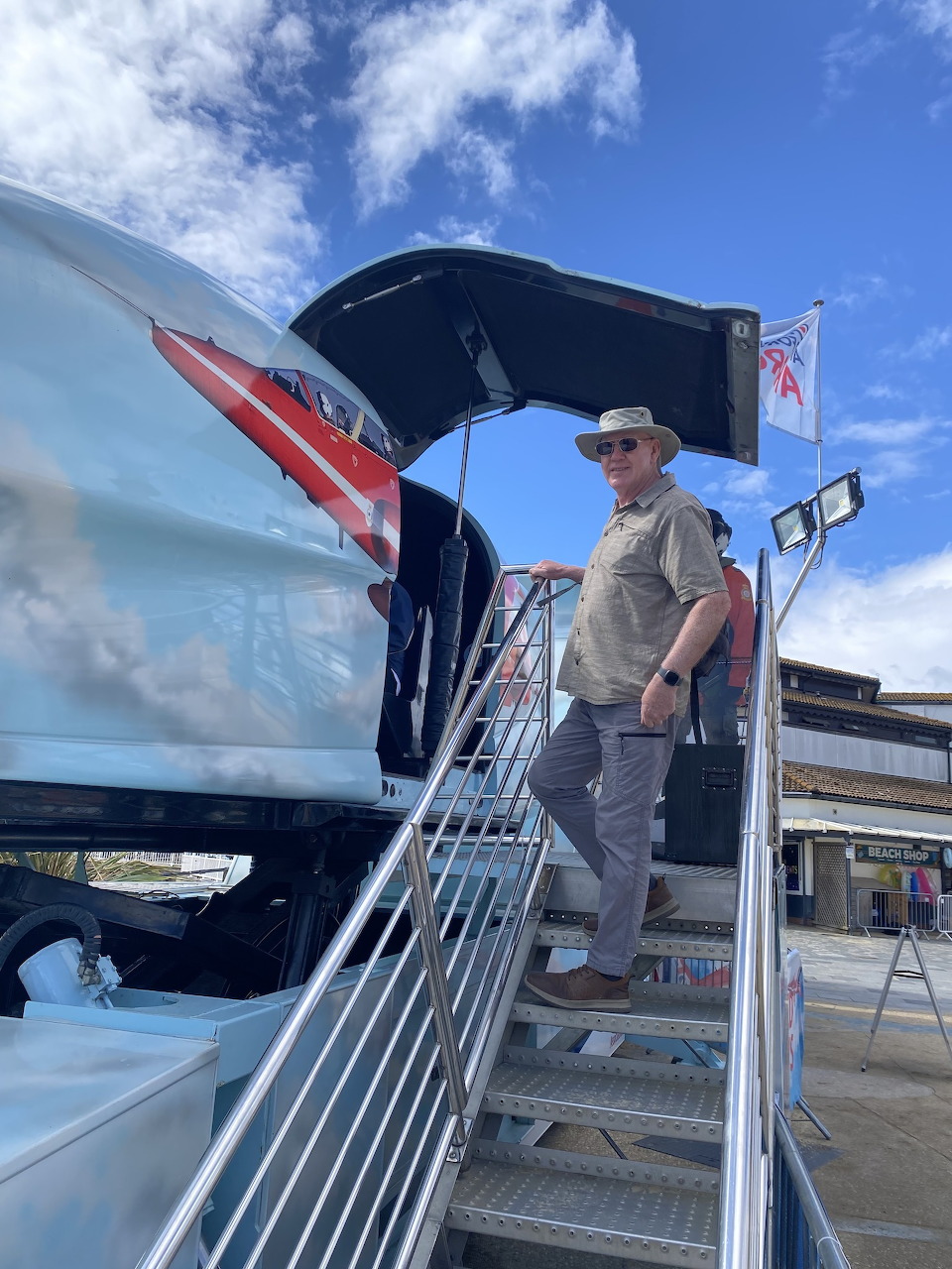 Man in hat on metal stairs next to a Red Arrows jet simulator cockpit outdoors