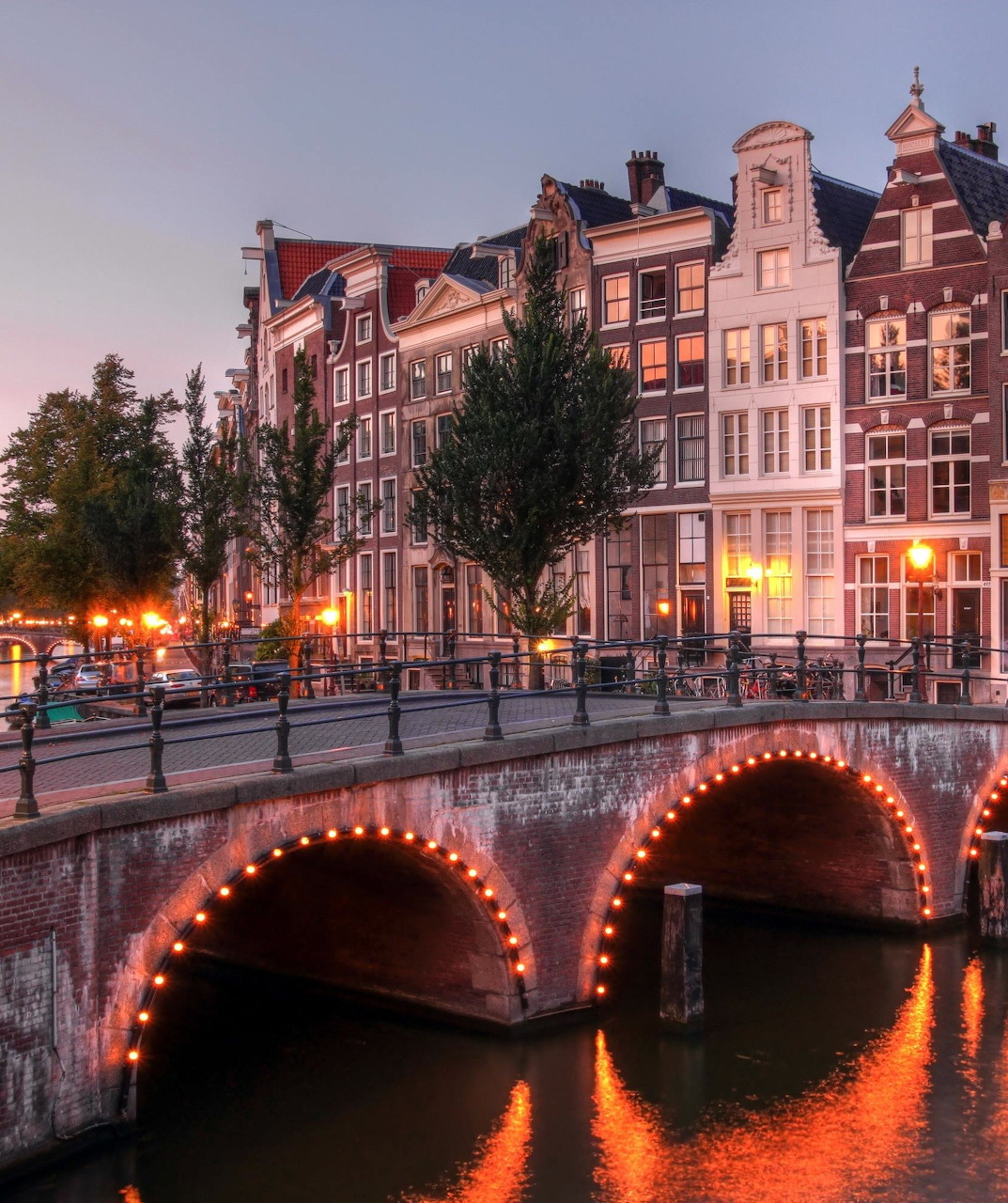 Illuminated canal bridge and traditional Dutch buildings reflected in water at dusk in Amsterdam