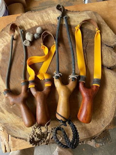 Four wooden slingshots with yellow rubber bands and small stones arranged on a rustic wooden board