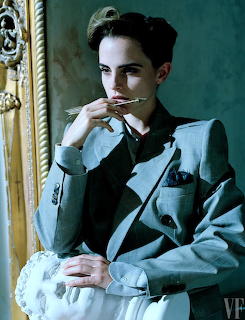 Portrait of Emma Watson in a gray suit, seated indoors with her hand near her face