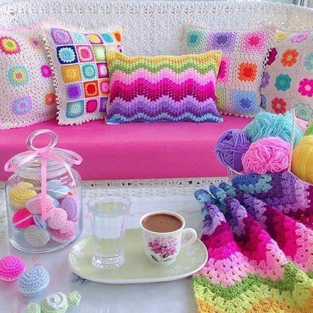 Colorful crochet pillows on a pink sofa with yarn balls, a coffee cup, and crocheted blankets