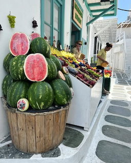 Barrel of watermelons at a fruit stand on a white-walled Greek island street with colorful fruits displayed