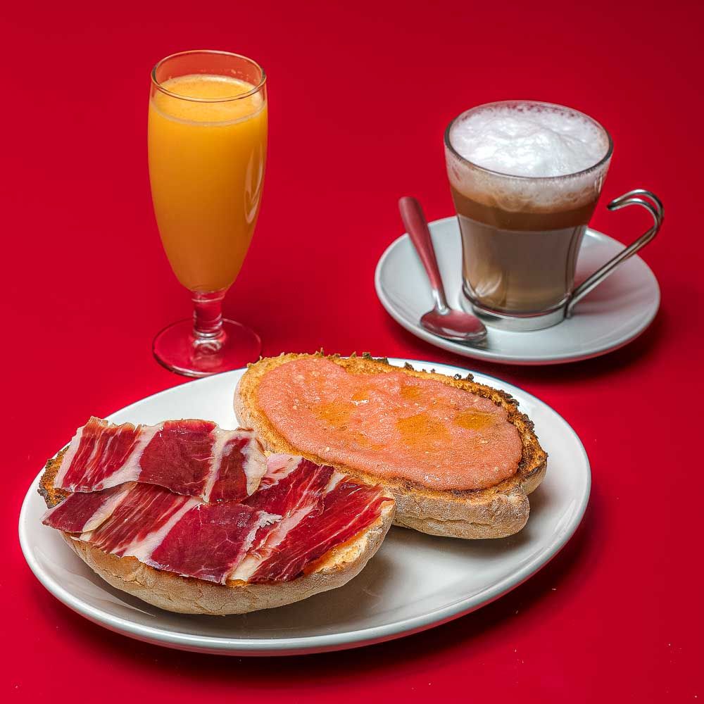 Spanish breakfast with ham on bread, tomato toast, glass of orange juice, and cappuccino on red background
