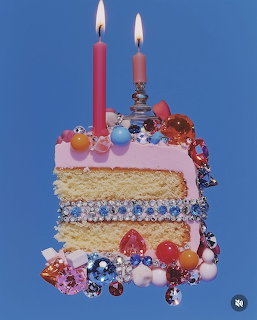 Slice of pink frosted cake with two lit candles and colorful gem decorations on a blue background