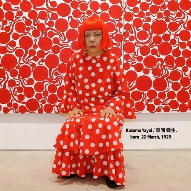 Yayoi Kusama sitting in a red polka dot dress in front of red and white abstract paintings