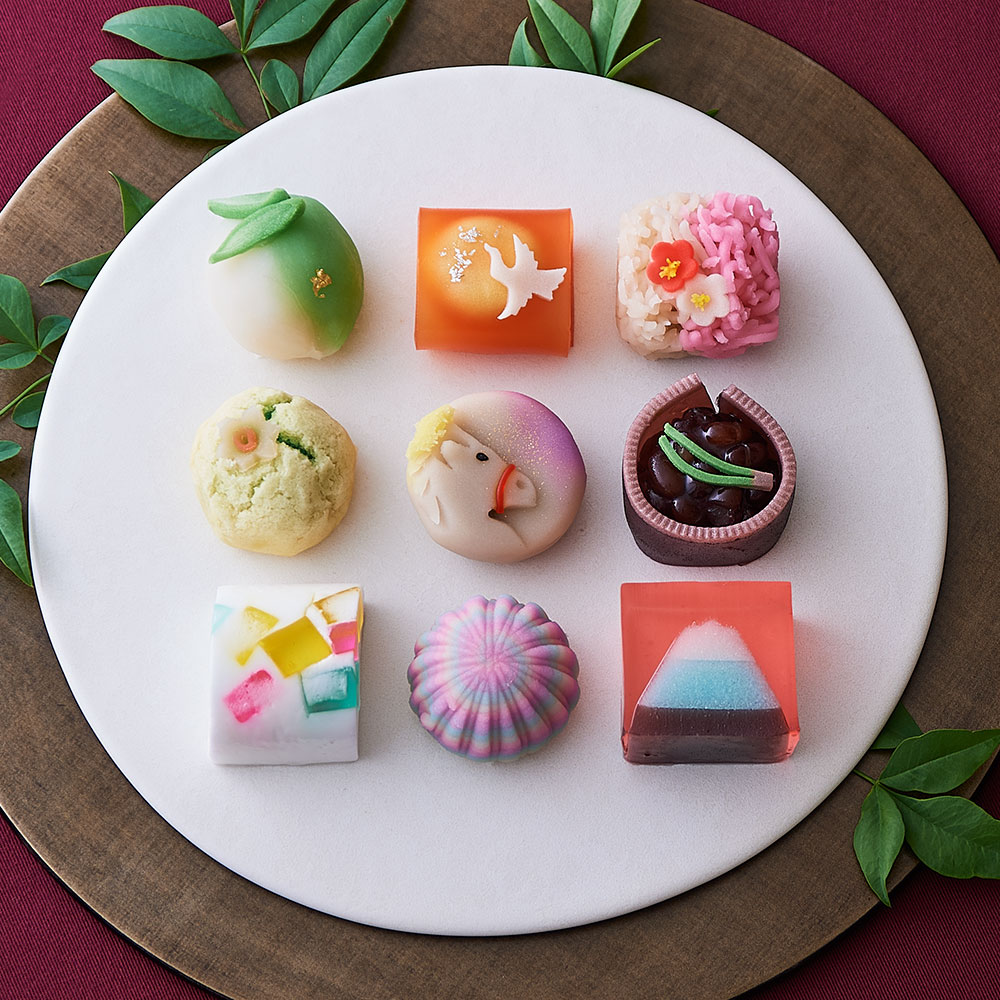 Nine decorative Japanese wagashi sweets arranged in a grid on a white plate with green leaves