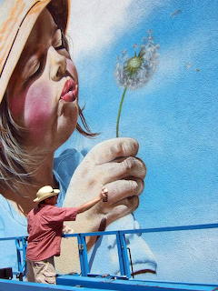 Street mural of a girl blowing dandelion seeds with artist on blue scaffolding below