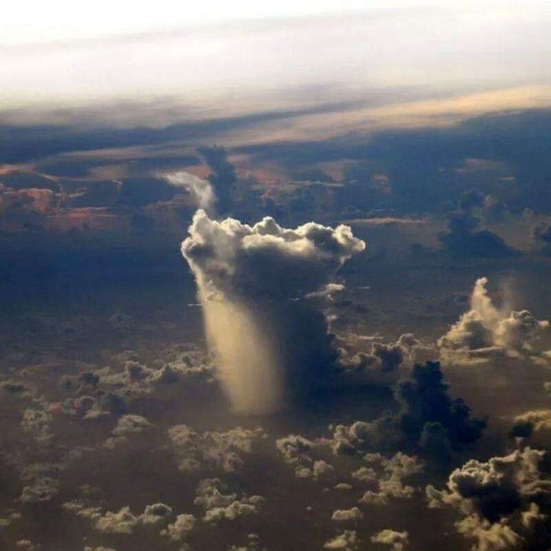 A towering cloud viewed from above with sunlight and a visible rain shaft below