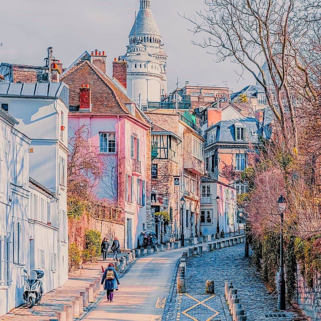 Pastel Paris street beside a narrow canal with bare trees and a round tower in the background