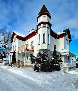 White Victorian house with a tower in snow beside a road under a blue sky