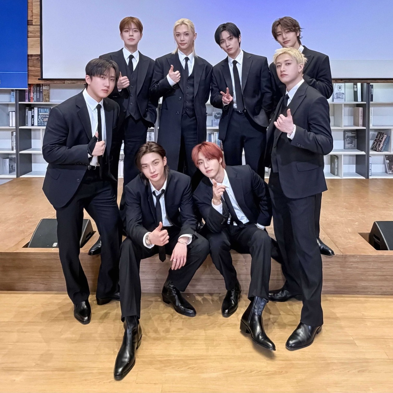 Eight Stray Kids members in black suits posing indoors in front of bookshelves