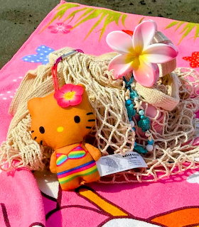 Hello Kitty toy with plumeria flowers and turquoise beads on a straw beach bag on colorful striped beach towel