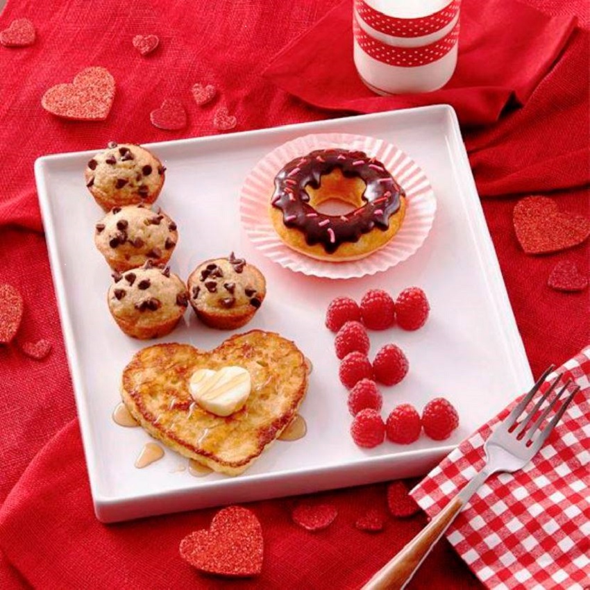 Valentine's breakfast with heart pancakes, donut, muffins, and raspberries on white plate with red hearts