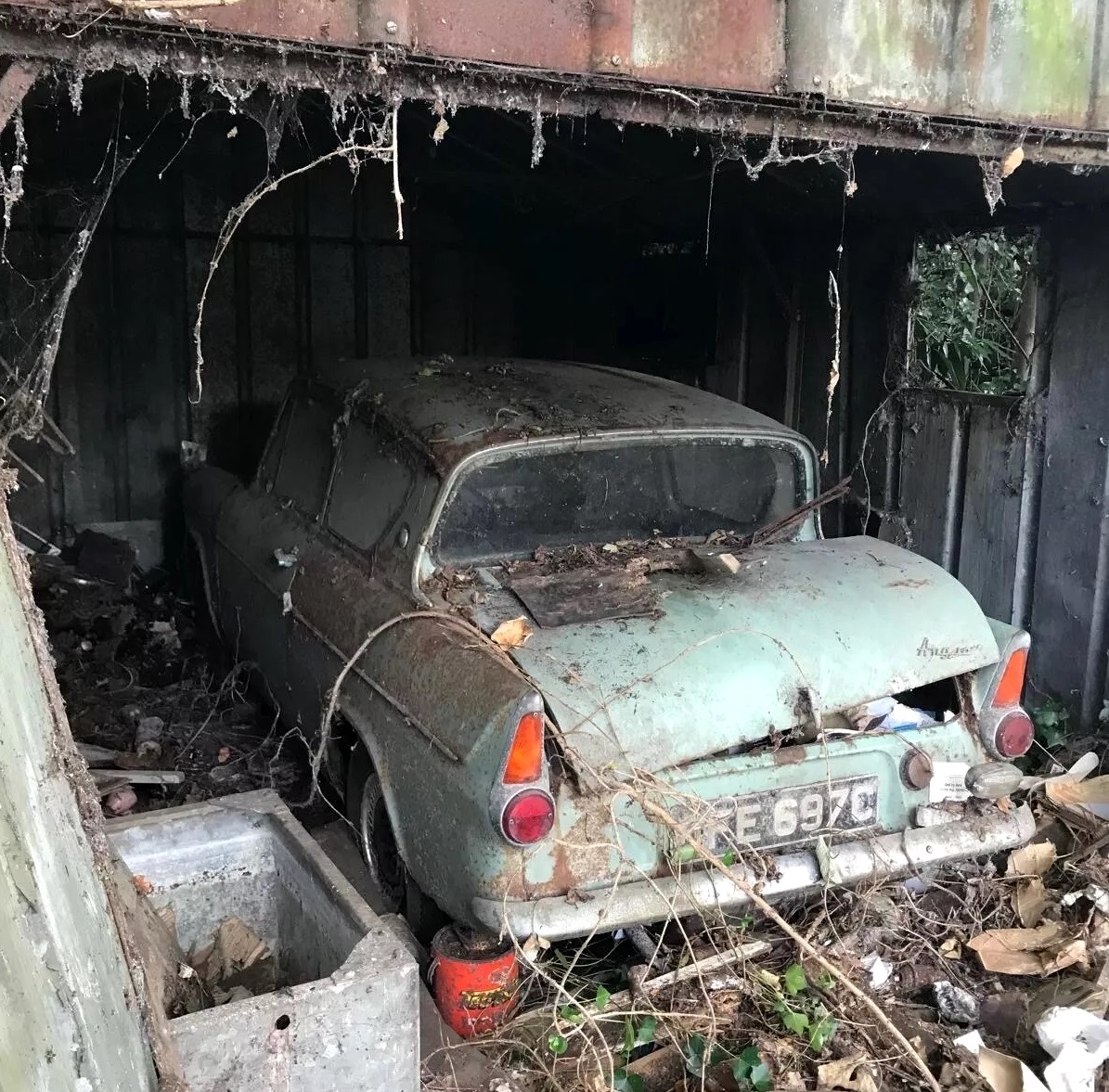 Rusted vintage Ford Anglia car abandoned in dilapidated building surrounded by vegetation and debris