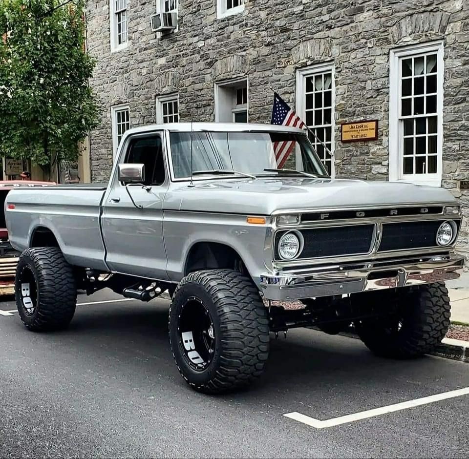 Silver lifted vintage pickup truck with large off-road tires parked beside a stone building