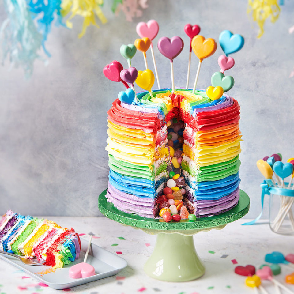 Colorful rainbow layered cake on a green stand topped with heart-shaped picks and filled with candy