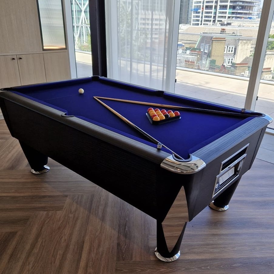 A black and blue pool table with pool cues and racked balls in a modern room with a city view.