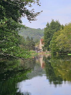 Calm pond reflecting trees and a church steeple surrounded by green hills