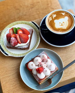 Coffee cup with bear latte art next to two small dessert plates with strawberries and cream on wooden table