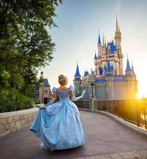 Woman in blue ball gown facing illuminated castle with blue spires at sunset