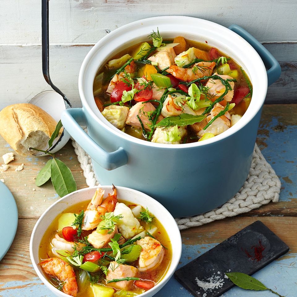Pot and bowl of fish stew with shrimp, vegetables, and fresh herbs on a wooden table