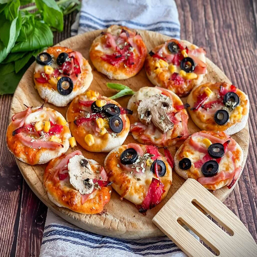 Assorted mini pizzas with various toppings on a round wooden cutting board with basil and a spatula