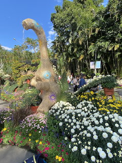 Large peacock topiary sculpture in garden with white chrysanthemums and colorful flowers