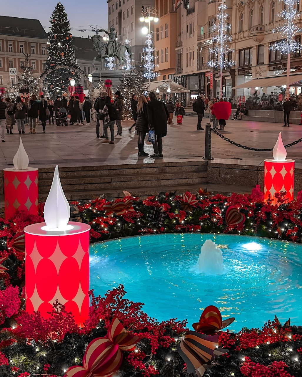 Zagreb Christmas market square at dusk with decorated fountain, red lanterns, Christmas tree, and people walking