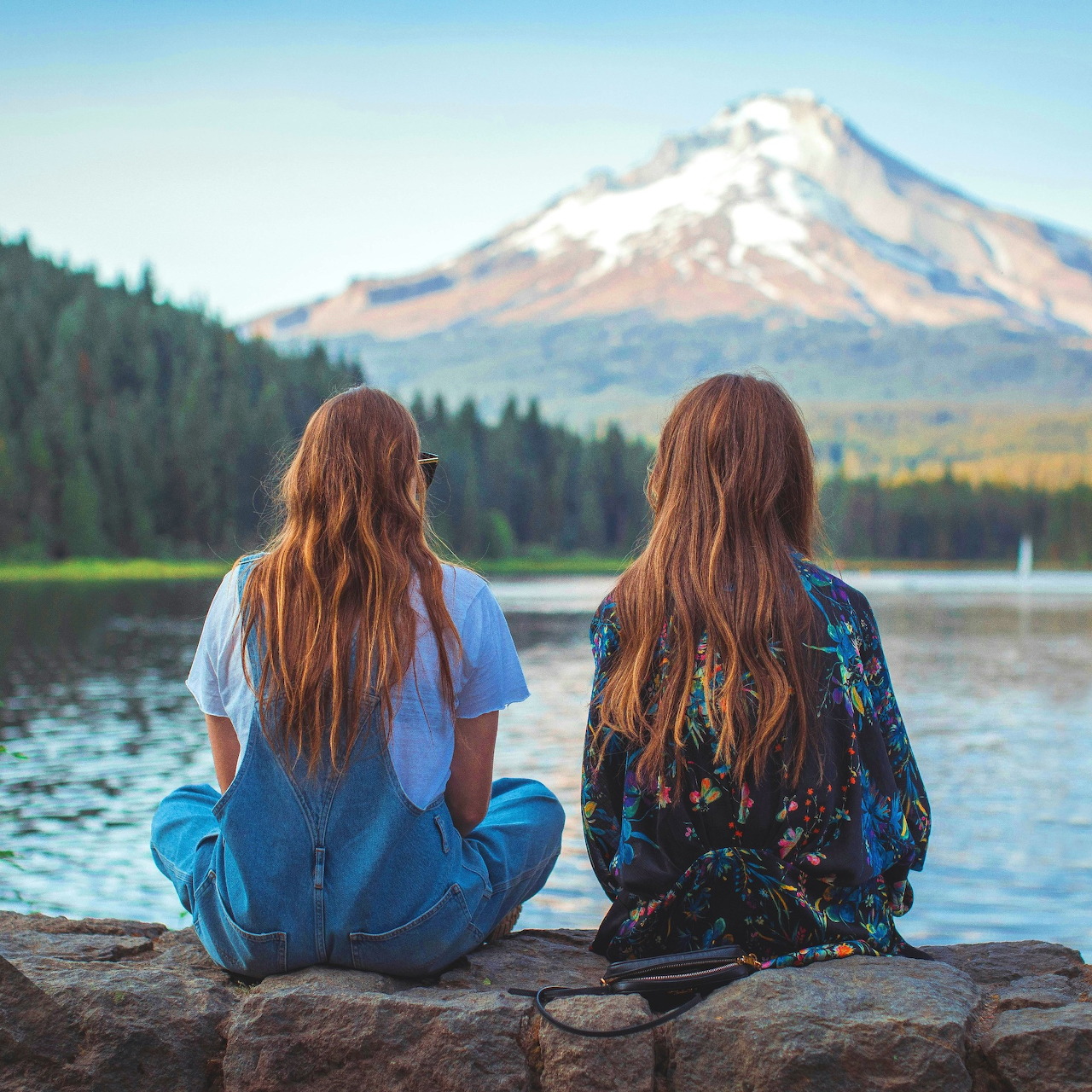 Two women with long red hair sitting by a lake looking at a snow-capped mountain