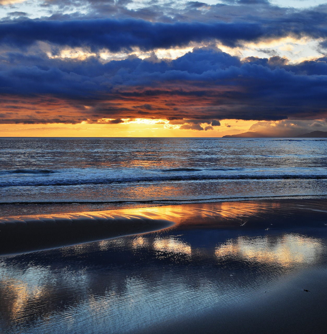 Sunset over the ocean with dark clouds and reflections on wet beach sand