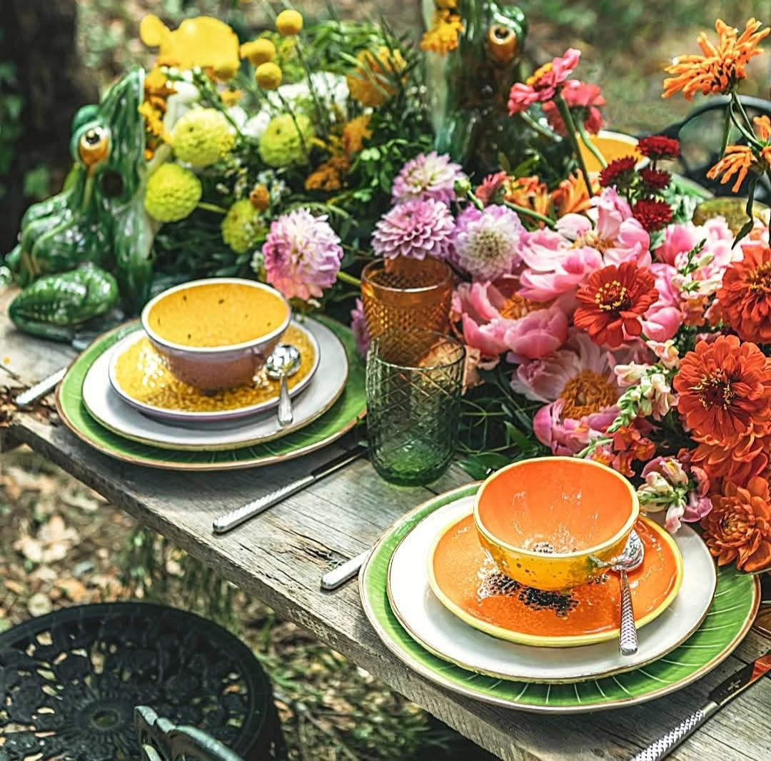Outdoor table with colorful flowers, orange and yellow plates, coffee cups on garden surface