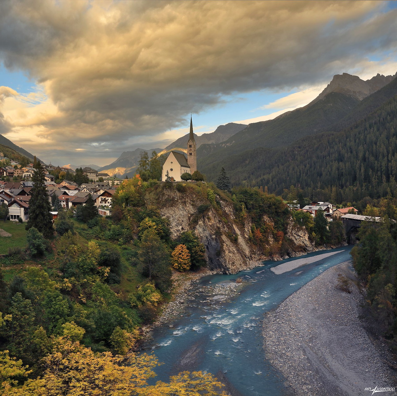 White church on rocky hill above turquoise river in mountain village with dramatic clouds